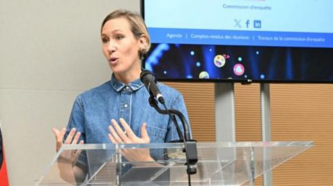 A woman in a denim shirt speaks at a lectern on the right while a man in a suit stands listening on her left