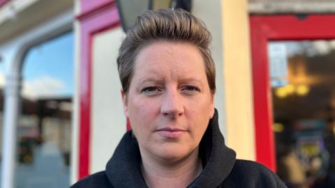 Sarah Godsoe is in the middle. She has short brown hair and is wearing a black jumper. She is stood in front of the outside of a pub.