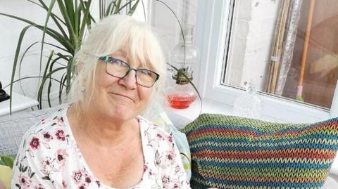 Lesley Lee with medium-length blond hair and glasses, smiling at the camera.  A multicoloured cushion is to her left and there is a spirit lamp with red liquid behind her, alongside a green pot plant.