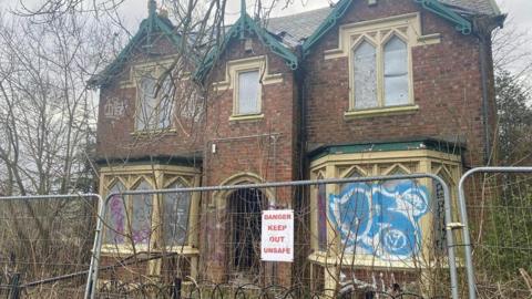 The Western Lodge which is a two-storey red brick building with big bay windows. The windows are boarded up with graffiti covering them. There's a fence surrounding the property with a 'DANGER KEEP OUT UNSAFE' sign.