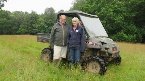 A man in a green fleece jumper and a woman in a dark blue fleece jumper standing next to a tractor on a farm.