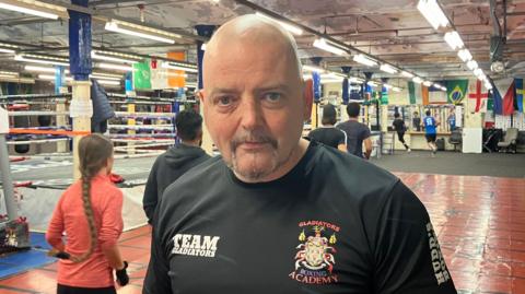 Den Doyle, a man with a grey goatee beard, stands in a boxing gym wearing a t-shirt saying Gladiators Boxing Academy. Behind him a number of people can be seen running around the room. Various colourful flags hand from the ceiling
