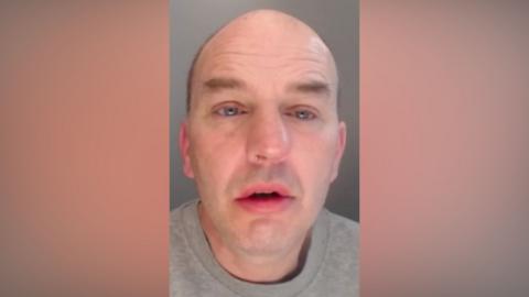 Neil Cook. He is bald and clean shaven and his eyes appear out of focus.
