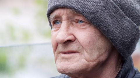 A close-up photo of a man's face. He is wearing a grey woollen hat and a black leather jacket. He has grey eyebrows and blue eyes.