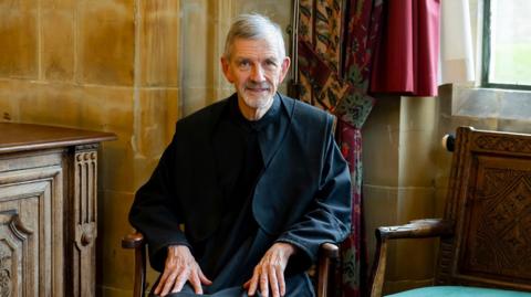 A man with short grey hair and facial hair wearing black robes. He is sat in a dark wooden chair with his hands on his lap