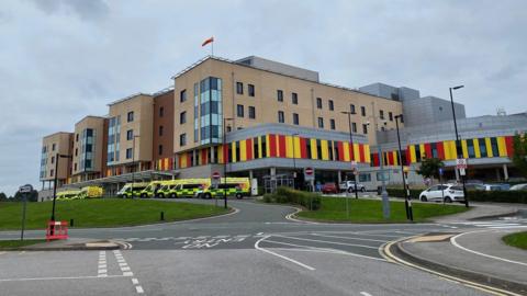 A hospital with ambulances parked outside it. The hospital building is several storeys high, and has red and yellow cladding on the lower part.