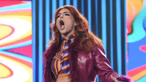 Jamie Hayter entering the arena for a wrestling match. She has long red hair, a maroon or purple jacket and ring gear that is purple, orange and red.