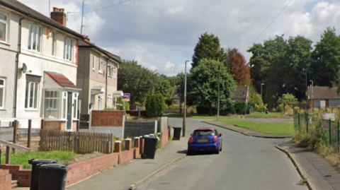 A google street view image of a residential street. Trees and a green space can be seen at the back of the road with houses lining the left hand side of the road