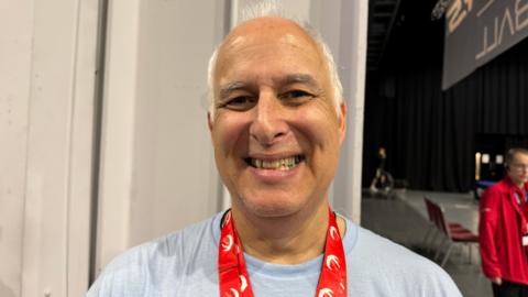 A man with white hair is smiling at the camera. He has a baby blue T-shirt on and a red lanyard with white logos on it
