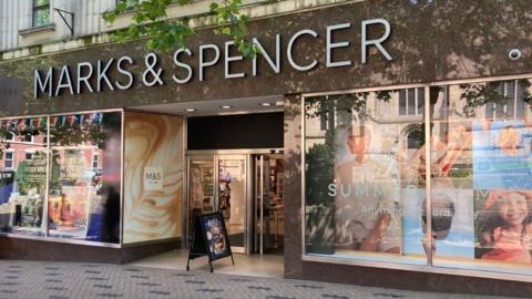 A shop front with a sign in white writing which says Marks & Spencer. There are colourful posters of people in the shop windows.