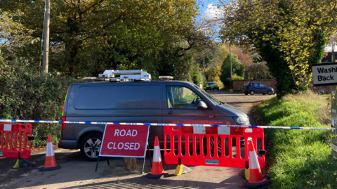 Part of Elm Lane in Copdock is closed off by blue and white police tape, a red 'road closed' sign, bright orange barriers and orange cones. A grey van is parked behind the cordon, it has been left across the road and would prevent any people or vehicles from getting past the road block. 