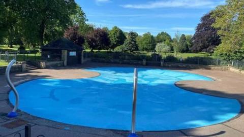 A children's paddling pool in a park, surrounded by a non-slip grey surface, with trees and play equipment in the background.