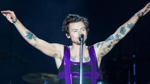 Harry Styles, who is wearing a sparkly purple and black top, has his arms in a V-shape in the air as he sings into a microphone. 