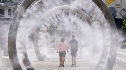 Two people holding hands walk through a water feature spraying a fine mist