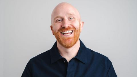 Ray Bradshaw - a bald man with a ginger beard, laughing as he looks into the camera. He is wearing a blue shirt.