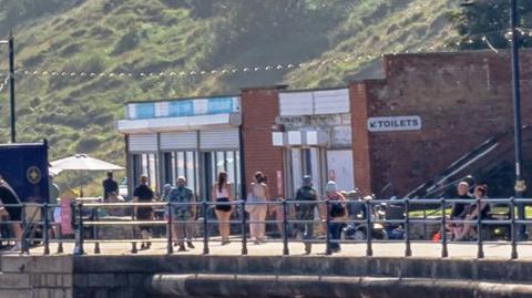 A small block with a sign reading "toilets" which is next to a fish and chip shop. It is above a beach with a hill behind it, and people are walking along the promenade in front of it.