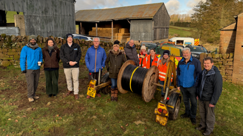 A group of residents wearing waterproof and outside clothing pictures at a rural location with barns in the background. There is a large roll of cable next to them. Some of those are wearing orange hi-vis clothing, suggesting they are part of the campaign to install the broadband. Everyone looks very happy.