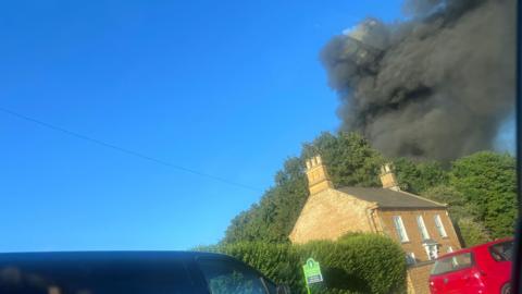 A thick pall of black smoke rises from behind trees. A van, car and cottage can be seen in the foreground.