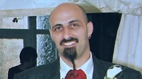 Napoleon Georgoulias is standing in front of a wall which has a mirror on the left. He has short hair and a long beard and moustache. He is wearing a white shirt, red tie and a grey blazer.