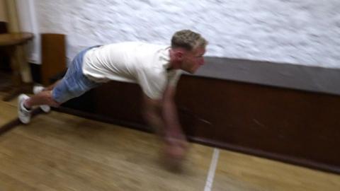 A man in jean shorts in mid-air throws a ball towards some skittles.