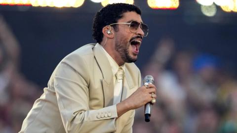 Bad Bunny in dark glasses, a beige suit, and cream shirt and tie singing into a microphone on stage during a concert in Barcelona on 22 May 2026