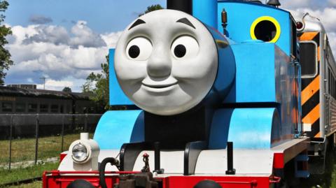 A replica of Thomas the Tank Engine. It is a blue model steam train with a grey smiley face on the front. 