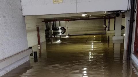 An underground car park is flooded with water. A car is at the back of the image and the water is up to its windows. A light is on in the middle of the ceiling. 