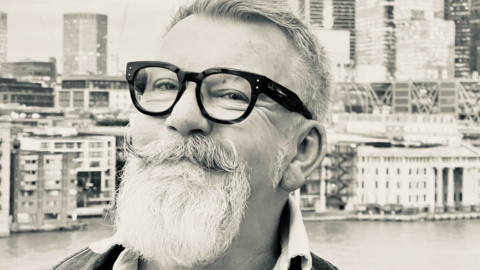 A black and white image of Simon Gee. He is smiling and wears dark-rimmed glasses. He has a distinctive moustache and beard. 