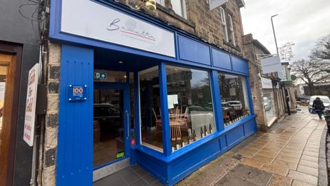 The store front of the restaurant Bavette. It is a royal blue colour, with two large windows at the front and a door on the left. Wine bottles and tables and chairs are visible through the windows. There is a white sign with the restaurant name above the door.