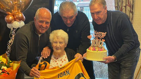 Steve Bull (l), Phil Parkes and Steve Daley are standing behind Joan who is sat in her chair smiling. They are holding a cake that says Happy 100th Birthday and a Wolves shirt with her name on across her lap. There's a bunch of orange and yellow flowers and gold and white balloons to the left side.