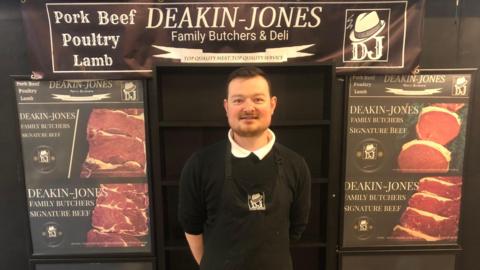 A man wearing a black top with a white collar and a black apron stands in front of a sign reading "Deakin-Jones Family Butchers & Deli". Other signs advertise the shop's "signature beef".