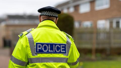 A stock image of a male police officer taken from behind to obscure his identity. He wears a hi-vis jacket and black hat.