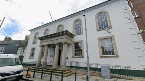 A courthouse with four pillars at the entrance. It's two floors. The windows at the bottom are square and the top windows have oval shapes at the top. It's cloudy. A white van is parked. A cone is on the pavement. 