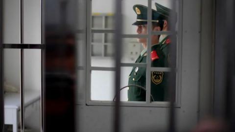 The bars of a prison cell. Through it you can see a man, who is of Chinese origin, wearing a military style uniform of China 

He looks stern and is possibly a prison guard