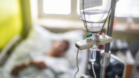 A image of a drip in the foreground in focus with a patient in a hospital bed in the background, out of focus