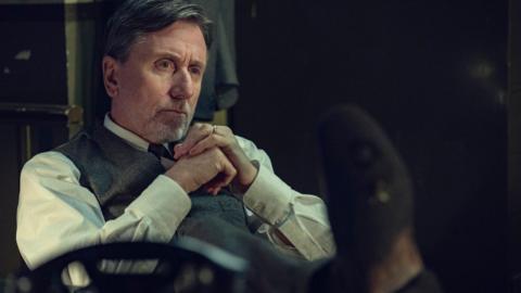 Actor Tim Roth is seated indoors, wearing a light shirt and a buttoned vest, with hands clasped near the chest. The setting appears dimly lit, with muted colours and blurred objects in the background suggesting an older or formal environment.