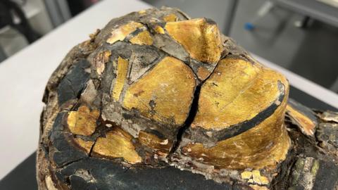 A close up of the gold funeral mask of Pa-Sheri.  There is a large crack across the cheek and around the nose of the mask.  Bits of the plaster have flacked away around the cracks.