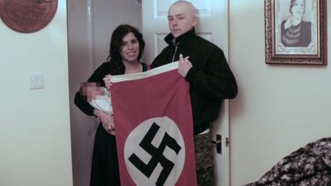 Claudia Patatas and Adam Thomas, holding their baby and a Swastika flag
