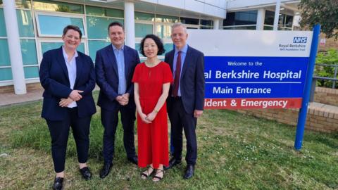 Two men wearing suites and two women, one wearing a suit and one wearing a red jumpsuit, standing next to a blue and white sign for Royal Berkshire Hospital and smiling.