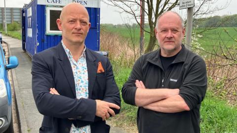 Two men with their arms folded stood a lay-by. A large green field, parking restriction sign and a blue-coloured cafe can all be seen in the background.