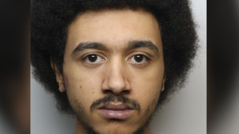 Custody photo of Jordan Crowley against a plain grey background. He has black facial hair on his cheeks and a moustache. He has dark brown eyes and afro hair. 