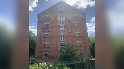 The outside of the former Surrey Advertiser building, which has the name of the paper on the front in black lettering. It is a six-storey mill building in red brick with windows all the way up and greenery growing around the ground floor.