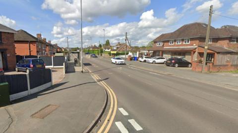 A Google street view image from a junction looking onto a main residential road. Cars are parked outside a building on the opposite side of the road, with a couple parked on the side of the road. 