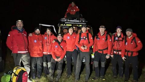 Nine of the rescuers stand in front of a land rover. Another team member is on top of the car. They all wear red jackets and black trousers. There are some backpacks in front of them on the left. It is dark. There are nine men and one woman in the team. 