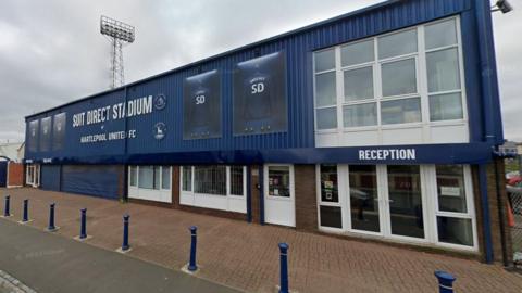 The Suit Direct stadium in Hartlepool, a blue two-storey structure with the reception area on the left. The stadium's floodlight can be seen towering in the background.