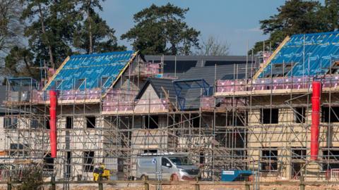 Scaffolding surrounds a couple of new build houses under construction. A silver van is parked outside. Three trees can be seen behind them on what is a sunny day.