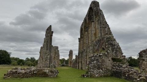 Ruined abbey walls stand on grass with some parts high and others lower, with the outlines of windows also seen. The sky is grey and cloudy.