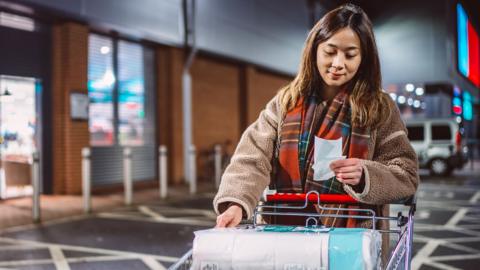 A woman stands outside a shop in a car park with a trolley containing loo roll and holding and looking down at a receipt.