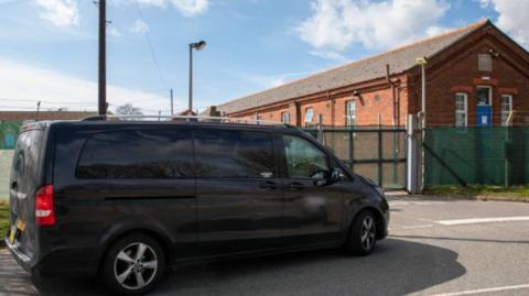 A black mini cab outside a centre for asylum seekers.  Its number plate and phone-number on its door are blurred