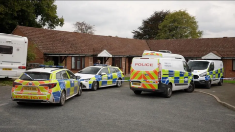 Two police cars and two police vans are parked outside the house, which appears to be a bungalow, with police tape surrounding them. The home, or homes, form an L shape in what looks like a cul de sac.
